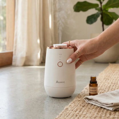 Hand holding a white Ausent diffuser with steam coming out, next to a bottle of essential oil on a textured surface.